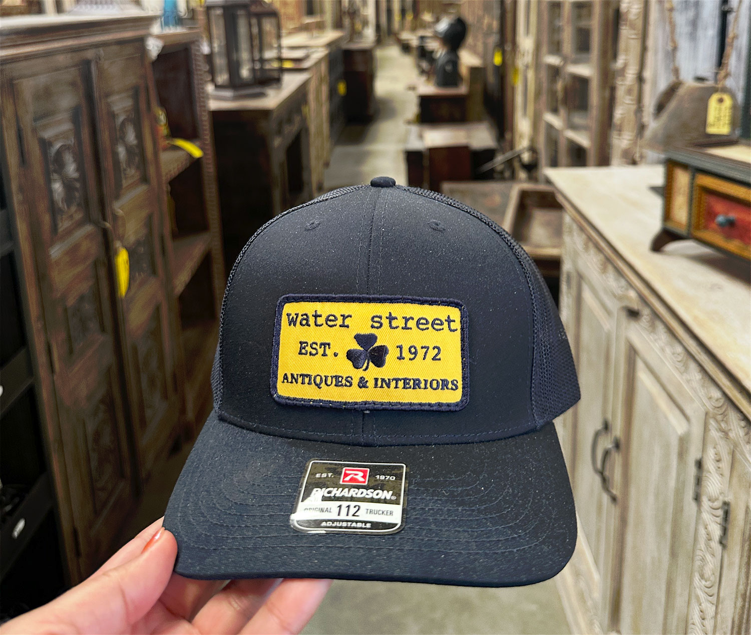 A trucker hat with a patch that wasy water street antiques since 1972 is held above a crowded warehouse of vintage furniture